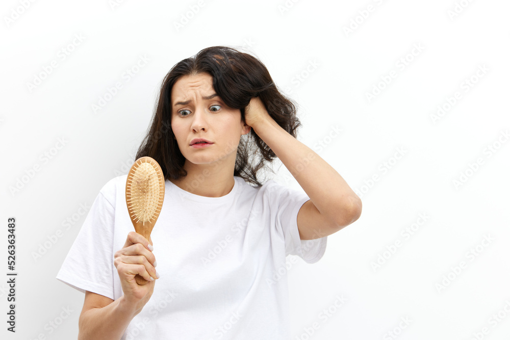 a sad, upset, puzzled woman with long black hair stands in a white T-shirt on a light background and holding her hand on her head in panic looks at her wooden comb a sad, upset, puzzled woman with long black hair stands in a white T-shirt on a light background and holding her hand on her head in panic looks at her wooden comb