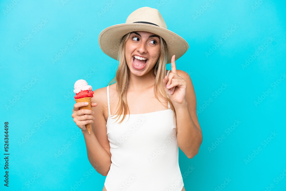 Young caucasian woman in swimsuit with a cornet ice cream isolated on blue background intending to realizes the solution while lifting a finger up Young caucasian woman in swimsuit with a cornet ice cream isolated on blue background intending to realizes the solution while lifting a finger up
