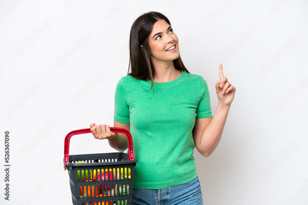 Young Brazilian woman holding a shopping basket full of food isolated on white background pointing up a great idea Young Brazilian woman holding a shopping basket full of food isolated on white background pointing up a great idea