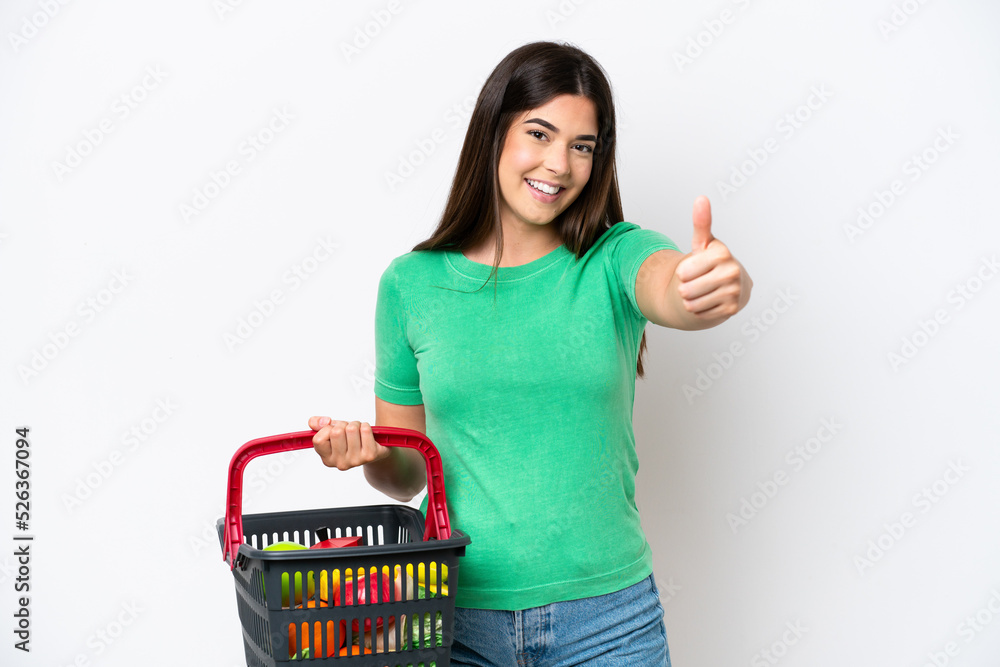 Young Brazilian woman holding a shopping basket full of food isolated on white background with thumbs up because something good has happened Young Brazilian woman holding a shopping basket full of food isolated on white background with thumbs up because something good has happened