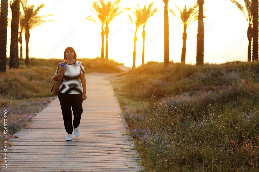 mature woman with sports mat is preparing to practice yoga outdoors on beach on sunrise. female is ready to do exercise on seashore on nature. copy space. Meditation, yoga and relaxation concept. mature woman with sports mat is preparing to practice yoga outdoors on beach on sunrise. female is ready to do exercise on seashore on nature. copy space. Meditation, yoga and relaxation concept.