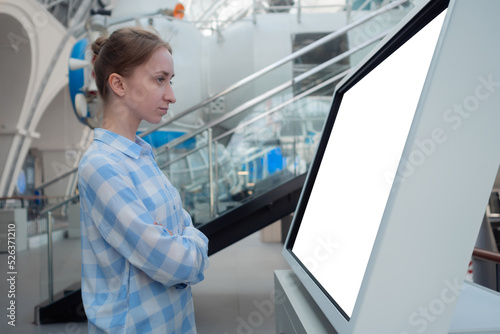 Woman in blue plaid shirt looking at blank interactive touchscreen white display of electronic kiosk at exhibition or museum: side view. White screen, mock up, copyspace, template, technology concept.