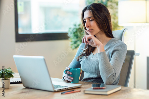 Concentrated beautiful business woman working with laptop while drinking coffee in living room at home.