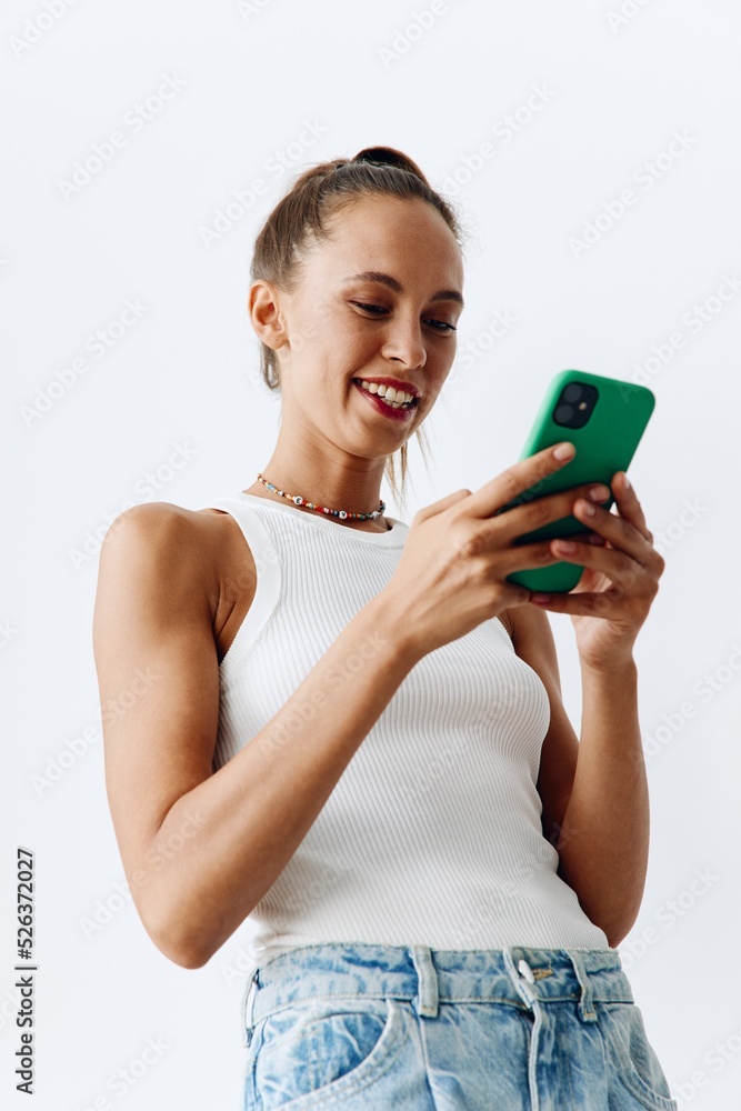 Millennial woman with beautiful and tanned skin looking at the phone screen in her hands and smiling in a white tank top and blue jeans against a white wall Millennial woman with beautiful and tanned skin looking at the phone screen in her hands and smiling in a white tank top and blue jeans against a white wall