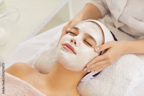 Close up of beautiful young girl with eyes closed enjoying white kaolin clay mask in beauty salon or spa center. Beautician applying calming cosmetic facial mask for fresh clear skin on woman's face