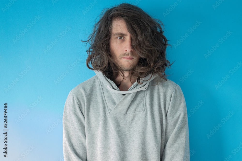 Serious long haired young man in grey hoodie on blue background Serious long haired young man in grey hoodie on blue background