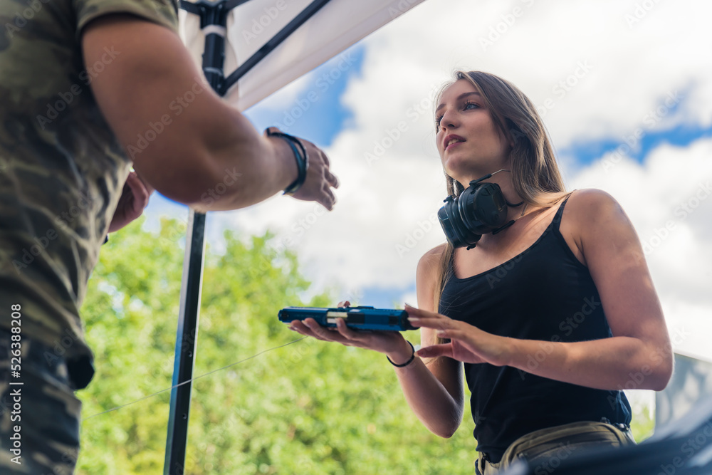 Naklejka premium Outdoor shot of gun reseller. Long-haired caucasian young adult girl in a black tank top holding a black gun and talking to the seller. Beautiful sunny weather. High quality photo