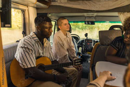 Multi-ethnic group of close friends on a summer road trip sitting inside their comfortable modern motorhome, playing the guitar, talking and laughing. High quality photo