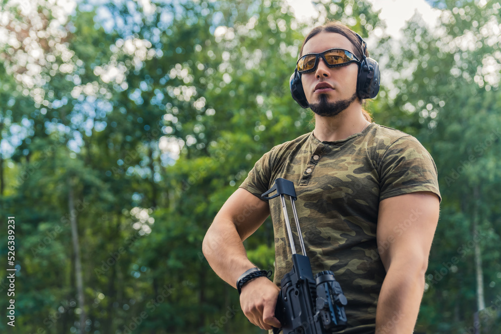 Medium outdoor shot of a military-looking caucasian muscular man in moro t-shirt looking far away at his target and holding black rifle with a loupe to look through. High quality photo