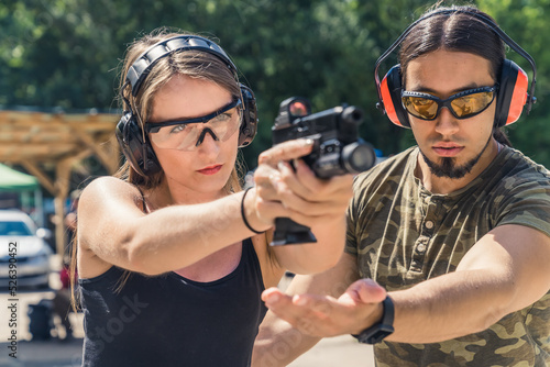 Outdoor gun range concept. Two caucasian adults - a man and a woman. Male instructor in protective gear in moro t-shirt helping out with his client's posture. High quality photo