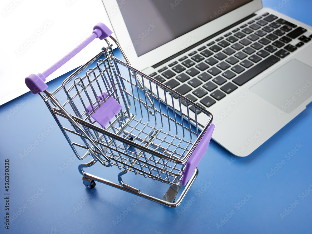 Small shopping cart and laptop on the blue table, close up. Toy grocery cart and notebook. The concept of electronic purchases, e-commerce.