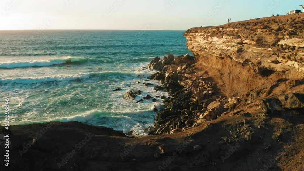 Rocky coastline alongside cliff, blue Atlantic Ocean water reflect at sunset. Nature landscape. Golden sandstone bluff beach. Tourists meet dusk. Travel destination. Fuerteventura Island, Canaries. Rocky coastline alongside cliff, blue Atlantic Ocean water reflect at sunset. Nature landscape. Golden sandstone bluff beach. Tourists meet dusk. Travel destination. Fuerteventura Island, Canaries.