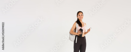 Full length of smiling healthy, slim asian girl going fitness training, female athelte carry backpack with workout equipment and water bottle, using mobile phone sports application, white background