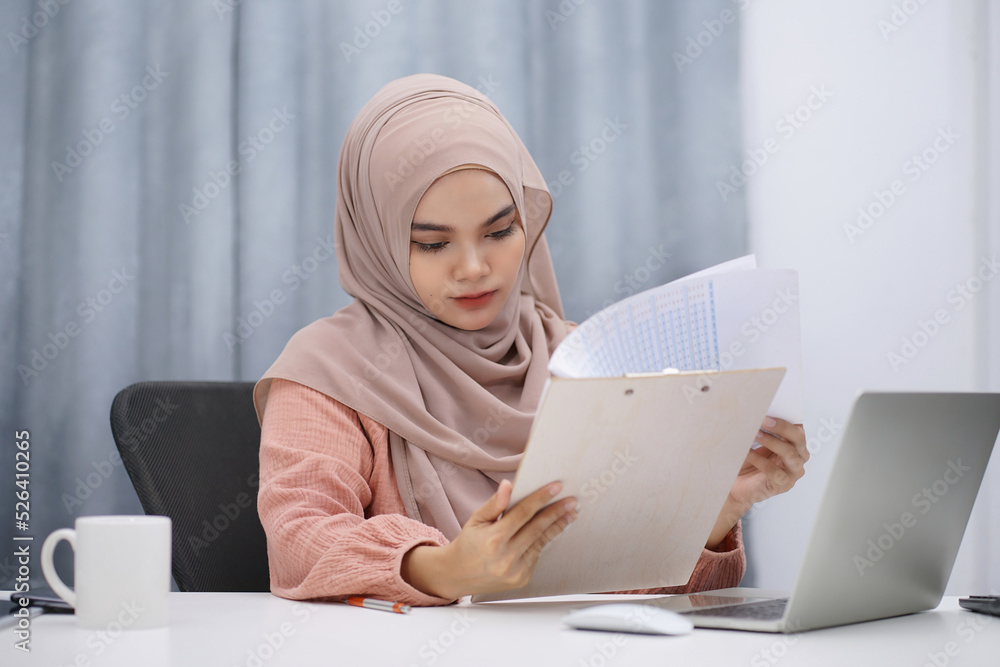 Happy asian muslim woman brown hijab working at home with laptop and documents. Happy asian muslim woman brown hijab working at home with laptop and documents.
