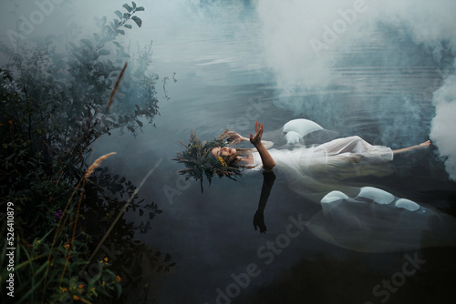 Fotografie, Obraz A lake nymph in a white dress and a wreath of flowers floats on the surface of the water