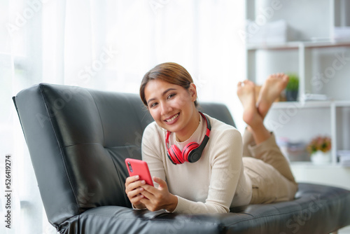 Pretty and cute Asian woman relaxing on the sofa, playing social media and accessing apps various entertainments, wear headphones, listen to music happily at home.