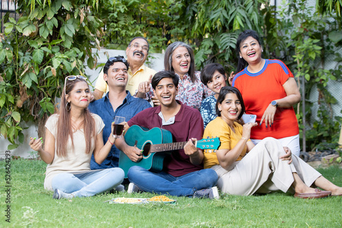 Portrait of happy multi generational indian family sitting enjoy picnic together outdoor garden playing guitar and music having fun., Summer party,. 