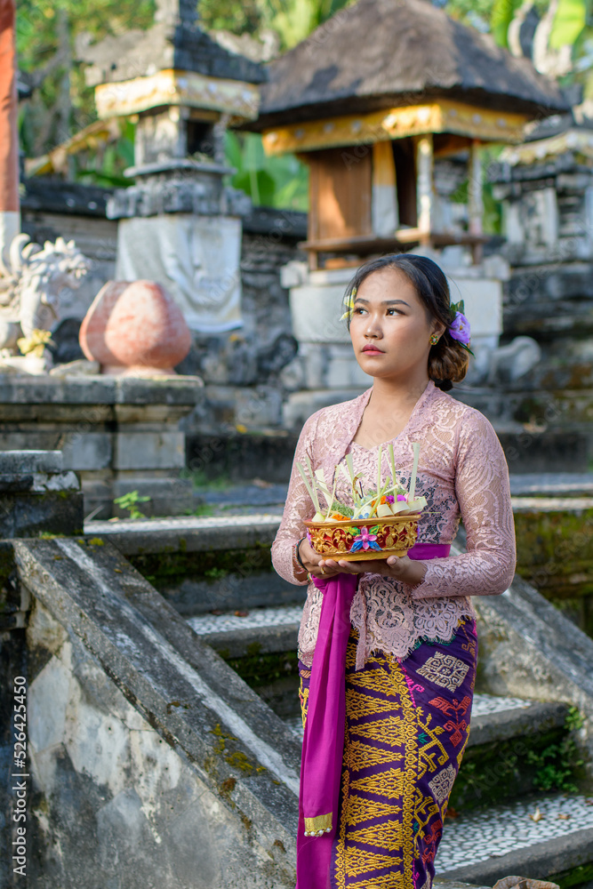 Beautiful Bali girl dressed in colorful batik sarong carrying offerings