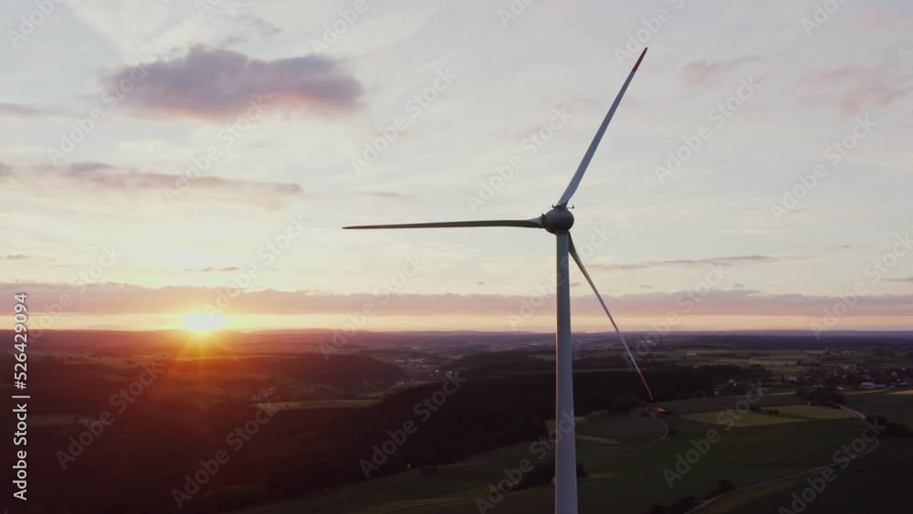 A close-up of the rotating blades of a wind turbine illuminated by the rays of the rising sun. Scenic green valley aerial view, drone video A close-up of the rotating blades of a wind turbine illuminated by the rays of the rising sun. Scenic green valley aerial view, drone video