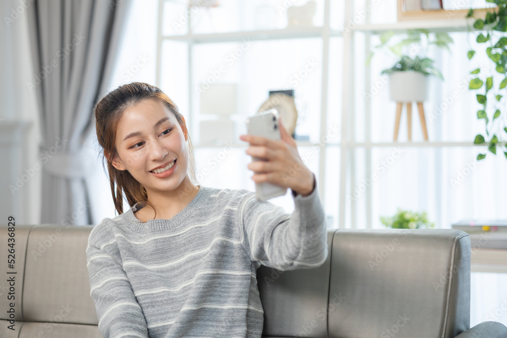 Happy and cheerful woman is using a phone sitting on a sofa.Asian young woman looking mobile phone for shopping online at home. Happy and cheerful woman is using a phone sitting on a sofa.Asian young woman looking mobile phone for shopping online at home.