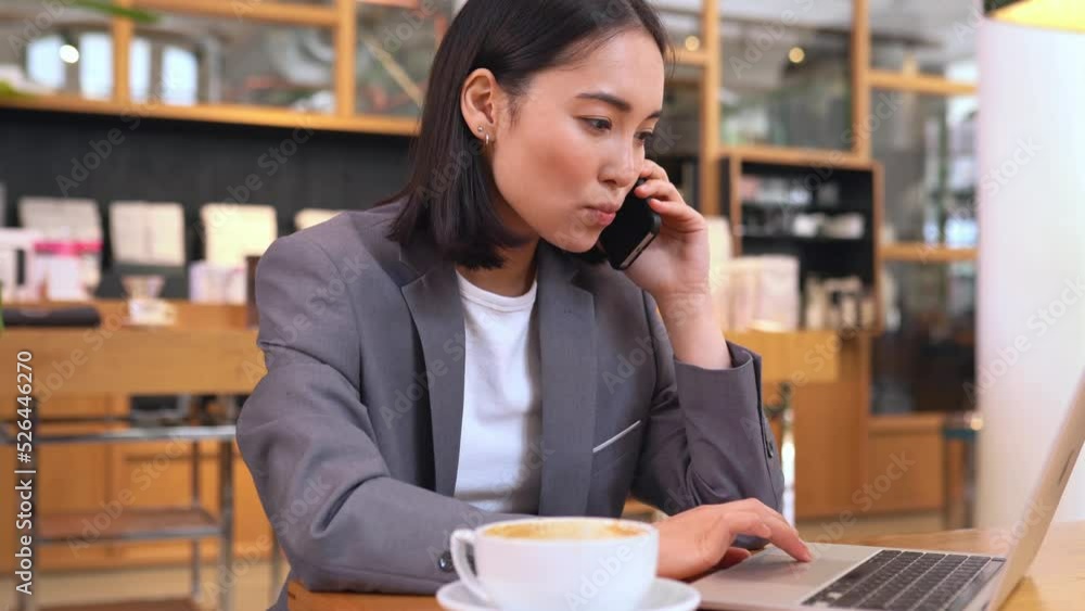 Smart smiling young Asian professional business woman using laptop talking on the phone having lunch in cafe. Korean lady entrepreneur or manager chatting on cell while remote distance working. Smart smiling young Asian professional business woman using laptop talking on the phone having lunch in cafe. Korean lady entrepreneur or manager chatting on cell while remote distance working.