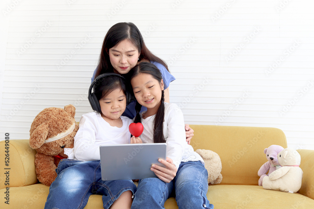 Adorable Asia happy family, cute two daughter girls with mother hold digital tablet, look and give red heart to web camera on video call, smiling mom and children have fun meeting online. Adorable Asia happy family, cute two daughter girls with mother hold digital tablet, look and give red heart to web camera on video call, smiling mom and children have fun meeting online.