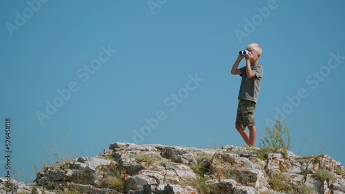 A fair-haired boy of seven years old, stands on the top of a cliff against the blue sky and looks somewhere into the distance around. He holds binoculars in his hands photo