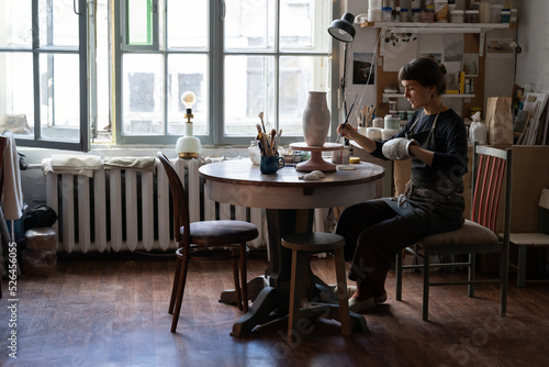 Female craftsperson in apron enjoys working with pottery craft in workshop. Young woman sits on chair at round table with equipment against bright window painting handmade ceramic vase with brush