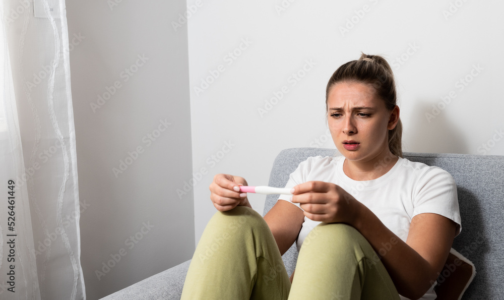 Young nervous woman is sitting at home holding a pregnancy test and is impatiently waiting for the test result. Maybe pregnant maybe not concept with anxious inpatient female on sofa in her apartment. Young nervous woman is sitting at home holding a pregnancy test and is impatiently waiting for the test result. Maybe pregnant maybe not concept with anxious inpatient female on sofa in her apartment.