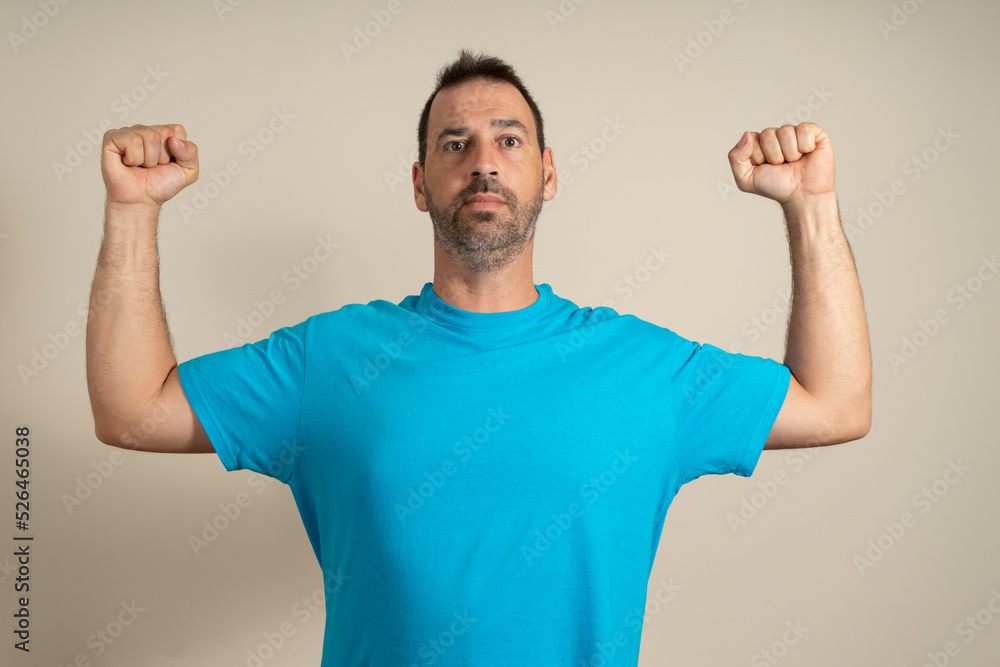 Fototapeta premium Caucasian happy unshaven strong sporty fitness student male 40s in casual blue t-shirt showing biceps muscles on hand isolated on beige background studio portrait