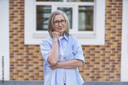 Mature grey hair woman wearing glasses standing outdoor traveling around world at retirement. Senior woman meeting her family. Mature woman with perfect skin wearing blue shirt standing outside