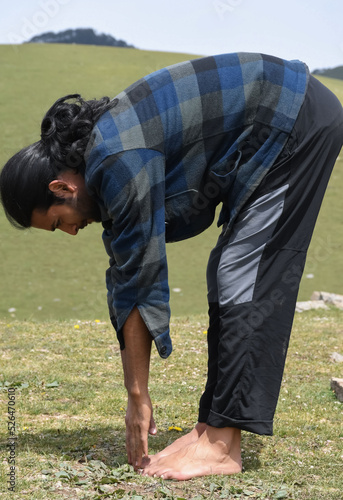 Side view of a long haired Indian young guy doing Uttanasana Yoga Pose in the mountain. A male hiker doing stretching by Standing Forward Bend Toe Touch exercise.