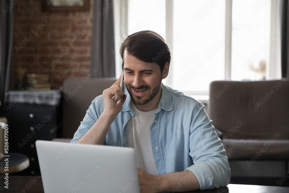 Happy engaged millennial freelance employee man answering cellphone call in home office, talking to client, giving phone consultation to customer on mobile phone, working at laptop computer, smiling Happy engaged millennial freelance employee man answering cellphone call in home office, talking to client, giving phone consultation to customer on mobile phone, working at laptop computer, smiling
