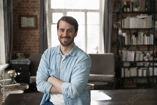 Happy confident millennial businessman in casual standing with arms crossed at work table in home office, looking at camera, smiling. Male freelance professional, business owner head shot portrait © fizkes