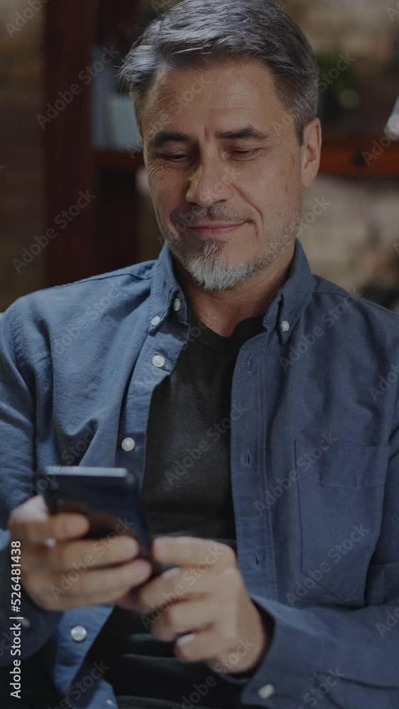 Older man at home, sitting on couch in living room, using phone, checking social media. Portrait of happy, mature age, middle age, mid adult caucasian man in 50s, smiling. Older man at home, sitting on couch in living room, using phone, checking social media. Portrait of happy, mature age, middle age, mid adult caucasian man in 50s, smiling.