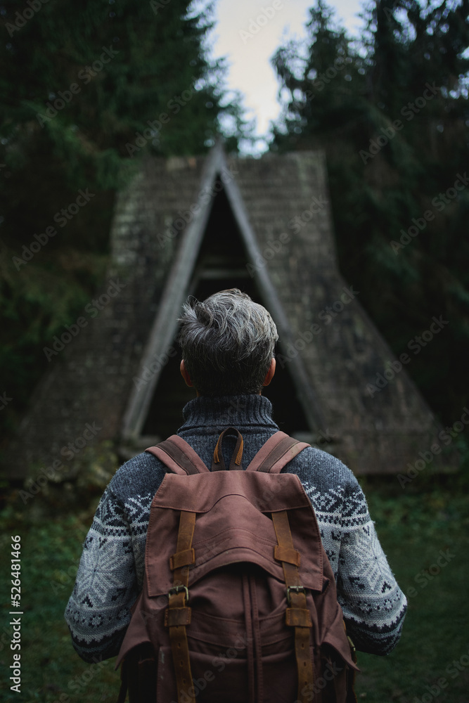 Vertical image back view traveler man with backpack standing in front of old vintage style wooden hut in the dark forest. scandinavian style moody aesthetic image