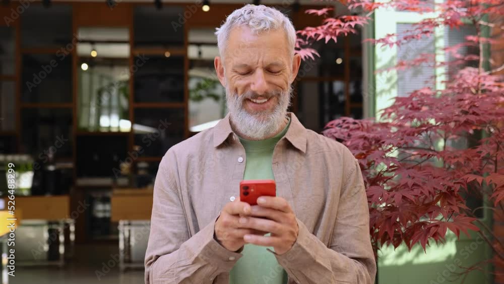 Smiling grey-haired older middle aged bearded man using moble phone outdoors. Happy old senior adult male user holding cellphone texting on smartphone, making bank payment or buying online. Smiling grey-haired older middle aged bearded man using moble phone outdoors. Happy old senior adult male user holding cellphone texting on smartphone, making bank payment or buying online.