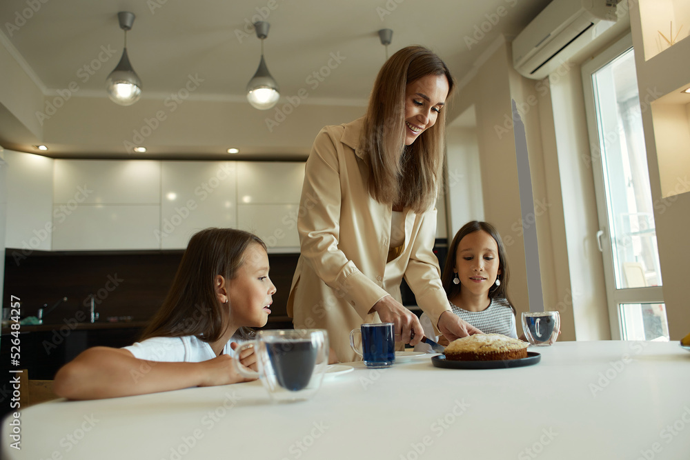 Charming attractive beautiful caucasian kind young smiling mother is drinking tea with her daughters, dressed in casual clothes, sitting in a bright kitchen, cutting pirok Charming attractive beautiful caucasian kind young smiling mother is drinking tea with her daughters, dressed in casual clothes, sitting in a bright kitchen, cutting pirok