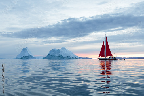 Sail boat with red sails cruising among ice bergs during dusk in front of a full moon. Disko Bay, Greenland. Midnight sun, romantic view. Climate change and global warming