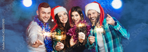 A group of young adult cheerful friends at a party wearing santa hats and tinsel with sparklers in their hands, laughing and looking at the camera on a dark smoky background. Banner for new year party