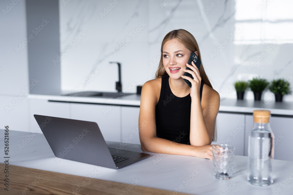 Young woman sits at the kitchen table using a laptop and talking on a phone Young woman sits at the kitchen table using a laptop and talking on a phone