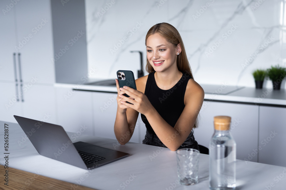 Joyful young woman using laptop and smartphone while sitting at kitchen. Joyful young woman using laptop and smartphone while sitting at kitchen.