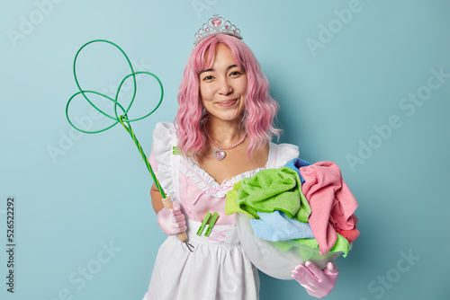 Pleased young Asian woman dressed like princess smiles pleasantly has long dyed pink hair holds basin of laundry and rug beater does domestic work isolated over blue background. Cleaning service photo