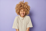 Portrait of good looking woman with curly hair laughs happily grins at camera keeps eyes closed expresses positive emotions dressed in casual t shirt isolated over purple background. Happiness