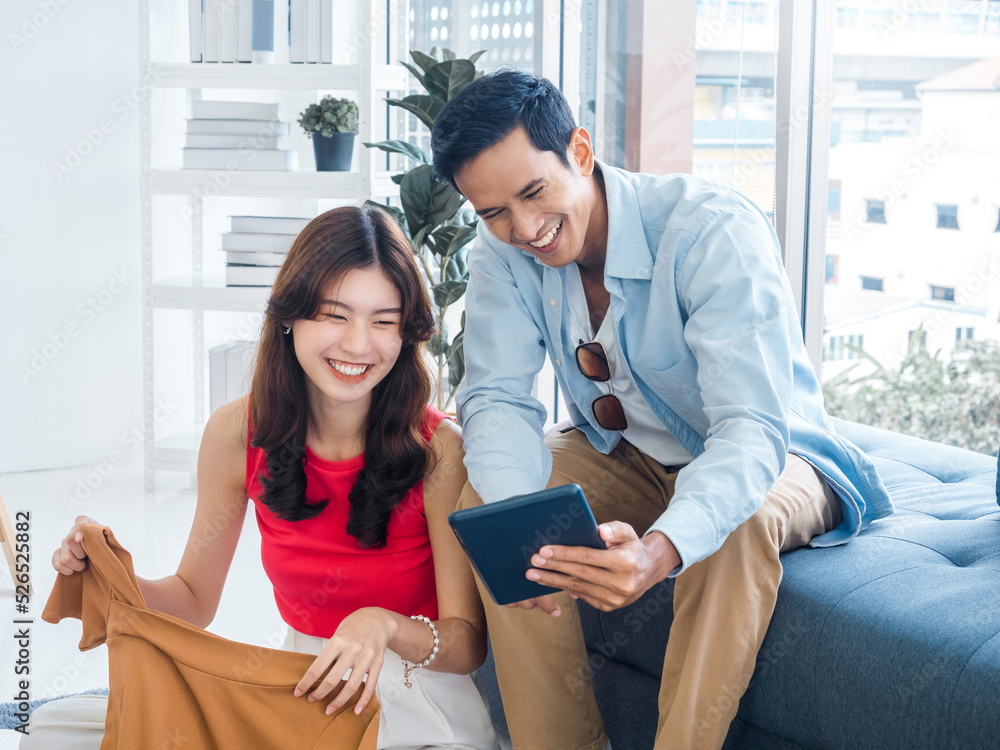 Happy handsome Asian man and young beautiful woman laughing together while looking at tablet screen. Woman hold a shirt and man holding tablet on couch near the glass window at white living room. Happy handsome Asian man and young beautiful woman laughing together while looking at tablet screen. Woman hold a shirt and man holding tablet on couch near the glass window at white living room.