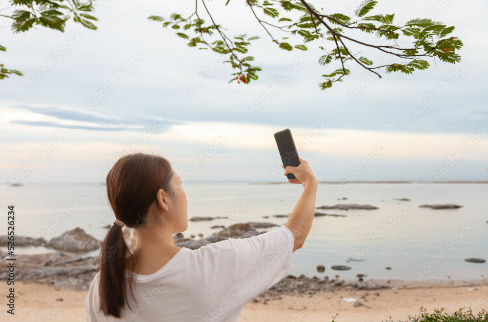 Asian women take photos and video calls at sea and beach in the morning at sunrise with their mobile phones, smartphones for posting to the internet online while traveling on vacation.
