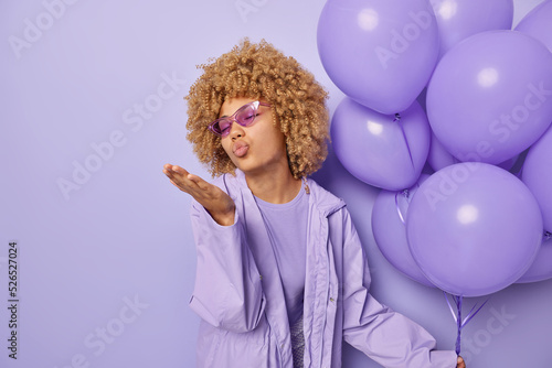 Fashionable curly haired woman blows air kiss expresses love wears suglasses and jacket holds bunch of inflated balloons celebrates special occasion isolated over purple background empty space