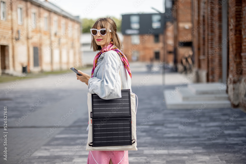 Fototapeta premium Young stylish woman walks with smart phone and charge it from portable solar panel hanging on bag. Concept of modern and sustainable lifestyle