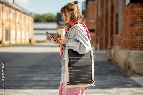 Young stylish woman walks with coffee and smart phone charging it from portable solar panel hanging on bag. Concept of modern and sustainable lifestyle photo