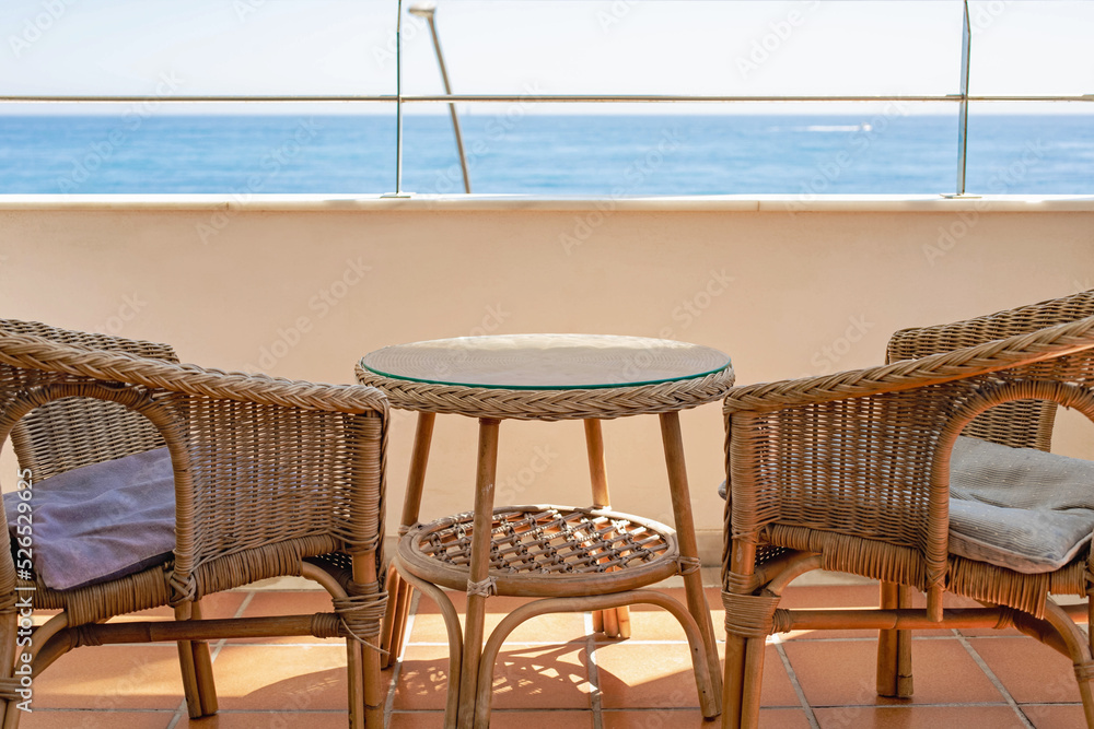Two wicker chairs and a round table are located on the terrace against the background of the blue sea.
